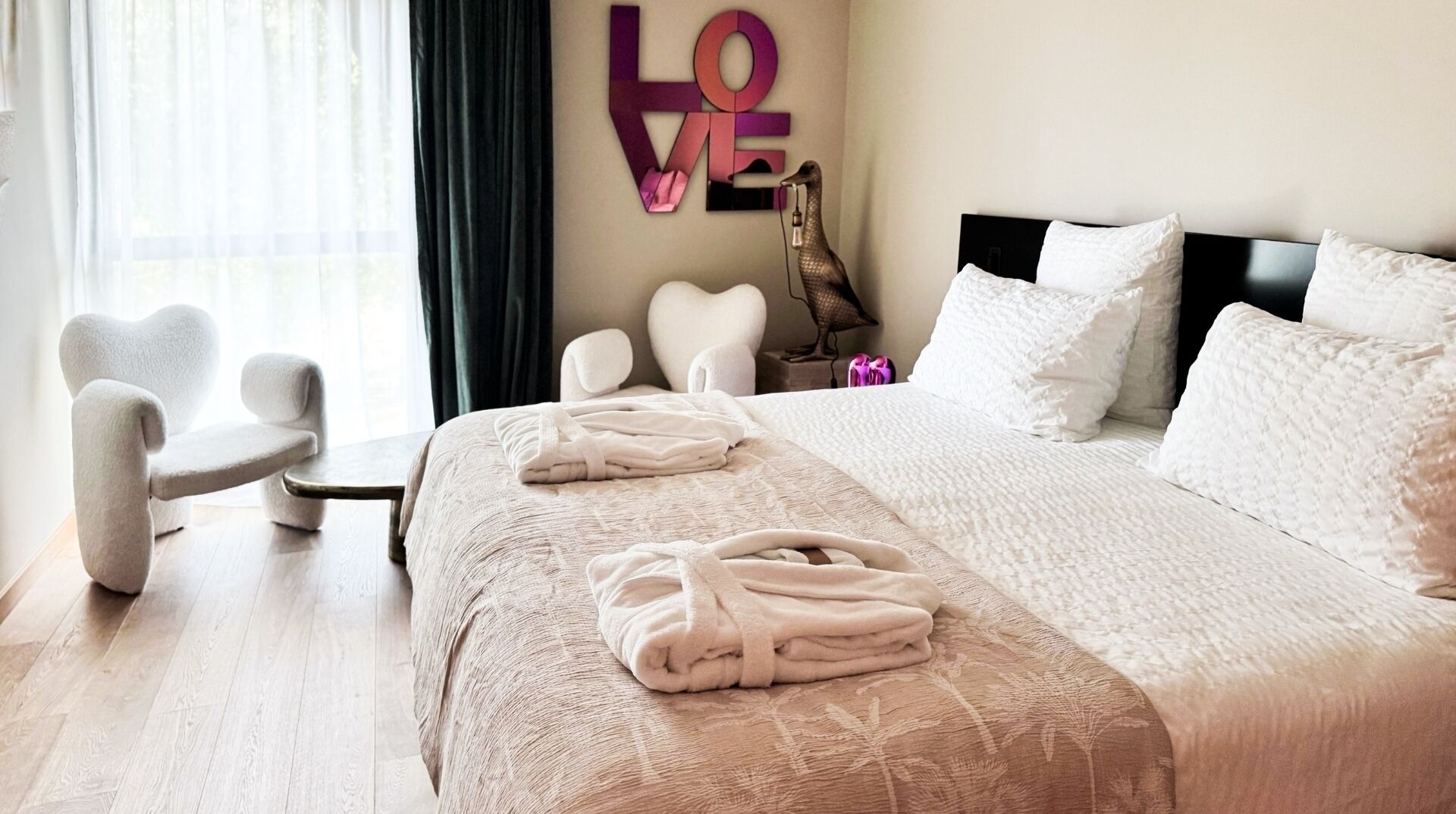 A hotel room with a large bed, a heart-shaped chair, a wall-mounted 'LOVE' sign, and a decorative bird sculpture.
