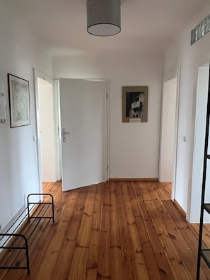 A room with wooden flooring, white walls, and a door leading to another room. There is a framed picture on the wall and a metal rack against the wall. - Apartment on Möser See-directly on the Havel cycle path, 5 minutes' walk from the beach (Brandenburg an der Havel)