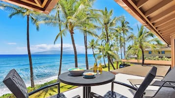 A table with two bowls and two bottles, overlooking a beach with palm trees and the ocean.