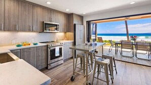 A modern kitchen with a large island, a microwave, and a refrigerator. There is a view of the beach through an open door.