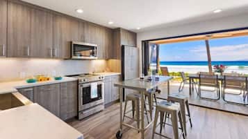 A modern kitchen with a large island, a microwave, and a refrigerator. There is a view of the beach through an open door.
