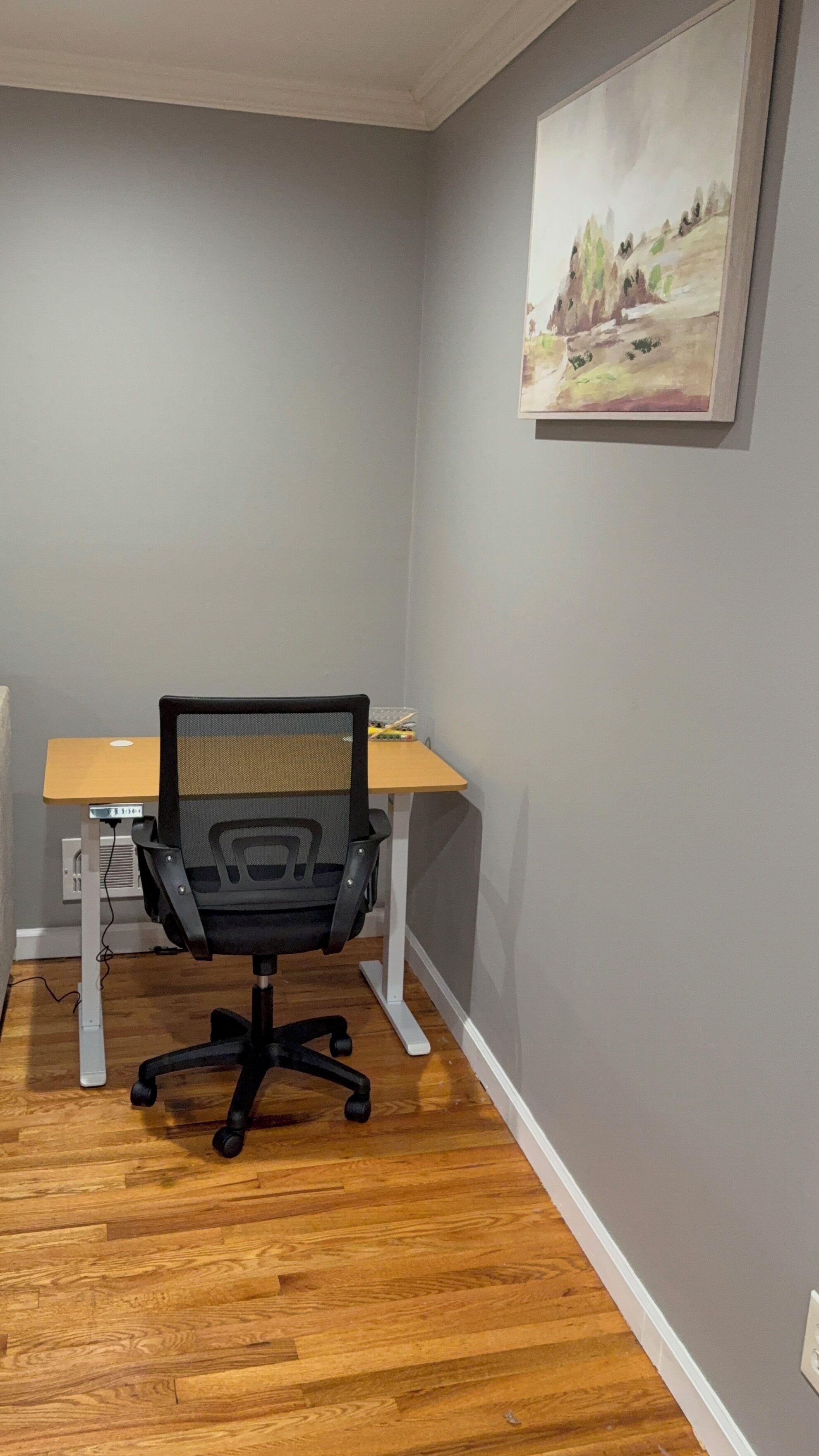 A small office space with a wooden desk, a black office chair, and a painting on the wall.