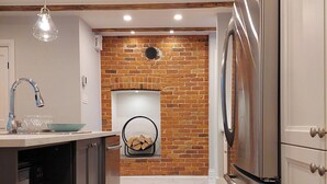 A kitchen with a brick wall, wooden flooring, a stainless steel refrigerator, and a hanging light fixture. - 25SV2-VE Ouimetoscope (Québec)