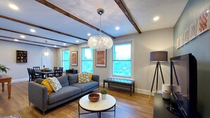 A spacious living room with a grey sofa, a coffee table, and a dining area with a wooden table and chairs. There is a large window providing natural light and a view of the outside. - 25SV2-VE Ouimetoscope (Québec)