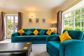 A room with a teal sofa set, yellow cushions, a dark coffee table, and a lamp. There are three framed pictures on the wall and a window with curtains. - Host & Stay | Alderfen View (Norwich)