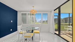 A bright dining area with a glass table and chairs, white walls, and large windows with blinds.