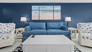 A living room with a blue sofa, two white wicker chairs, a white coffee table, and a beach-themed wall art.