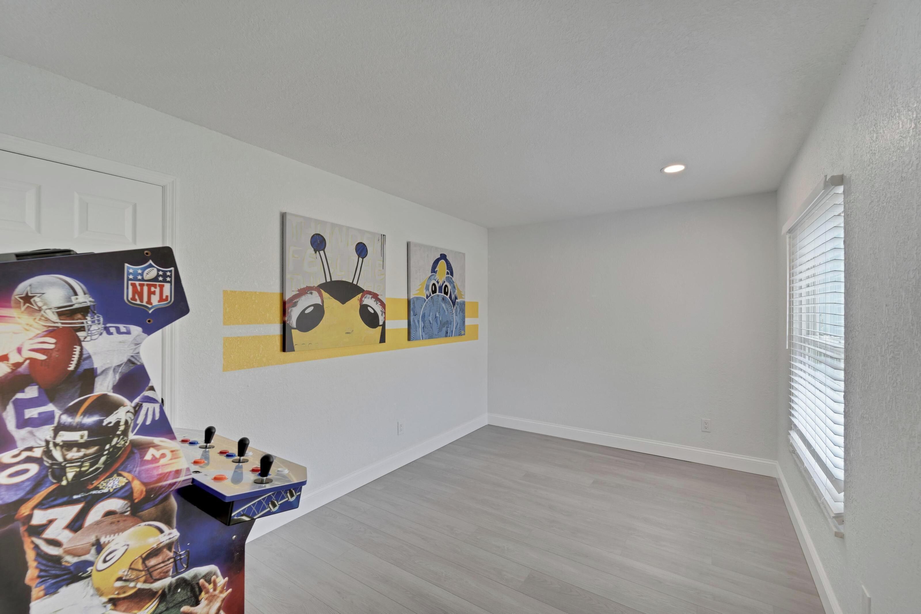 A room with a wooden floor, white walls, and a window with blinds. There is a wall-mounted air conditioning unit and a door.