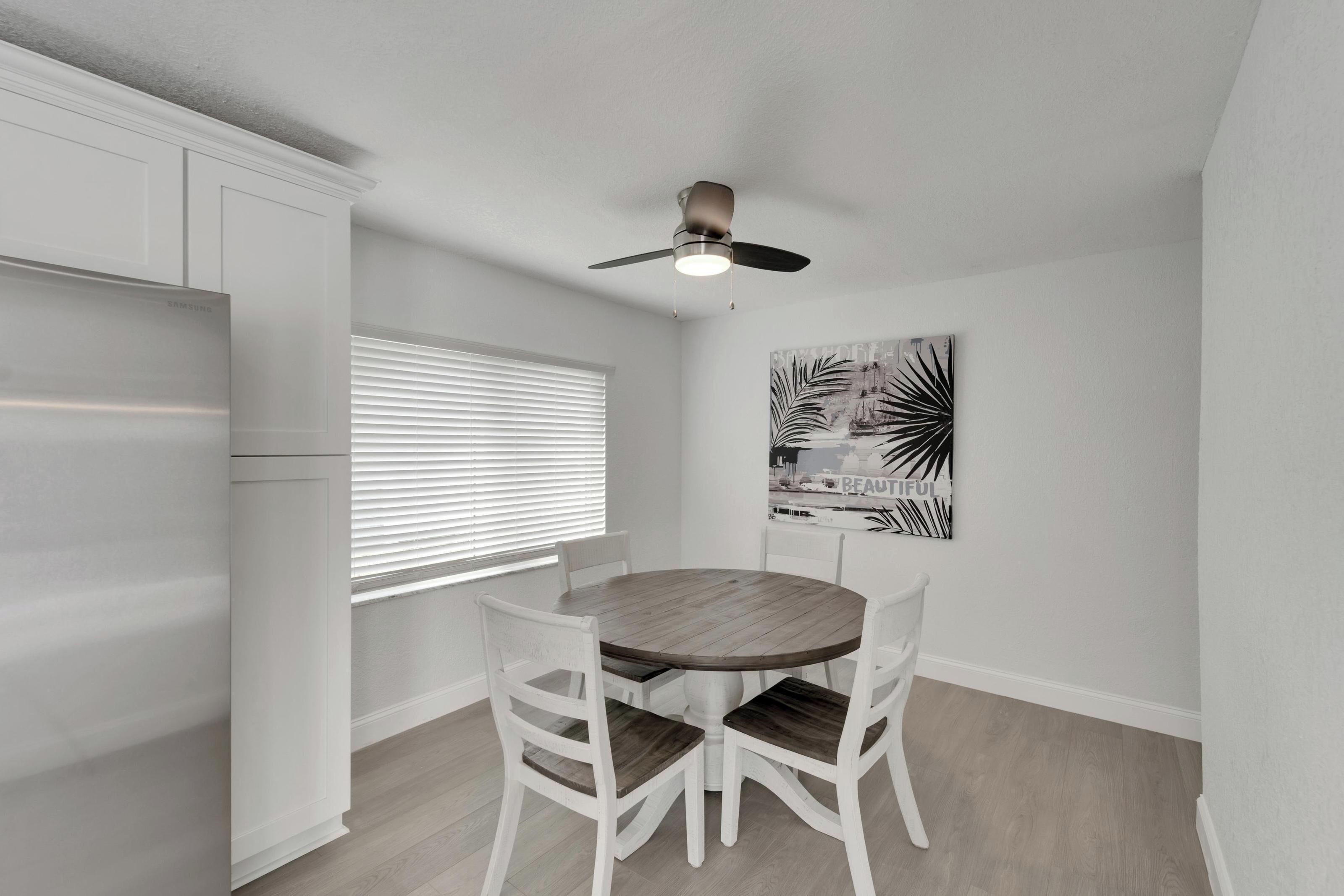 A dining area with a round table, four white chairs, a ceiling fan, and a window with blinds.