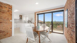 Een moderne keuken met witte kastjes, een eethoek met een houten tafel en witte stoelen, en een grote schuifpui naar een balkon met uitzicht.