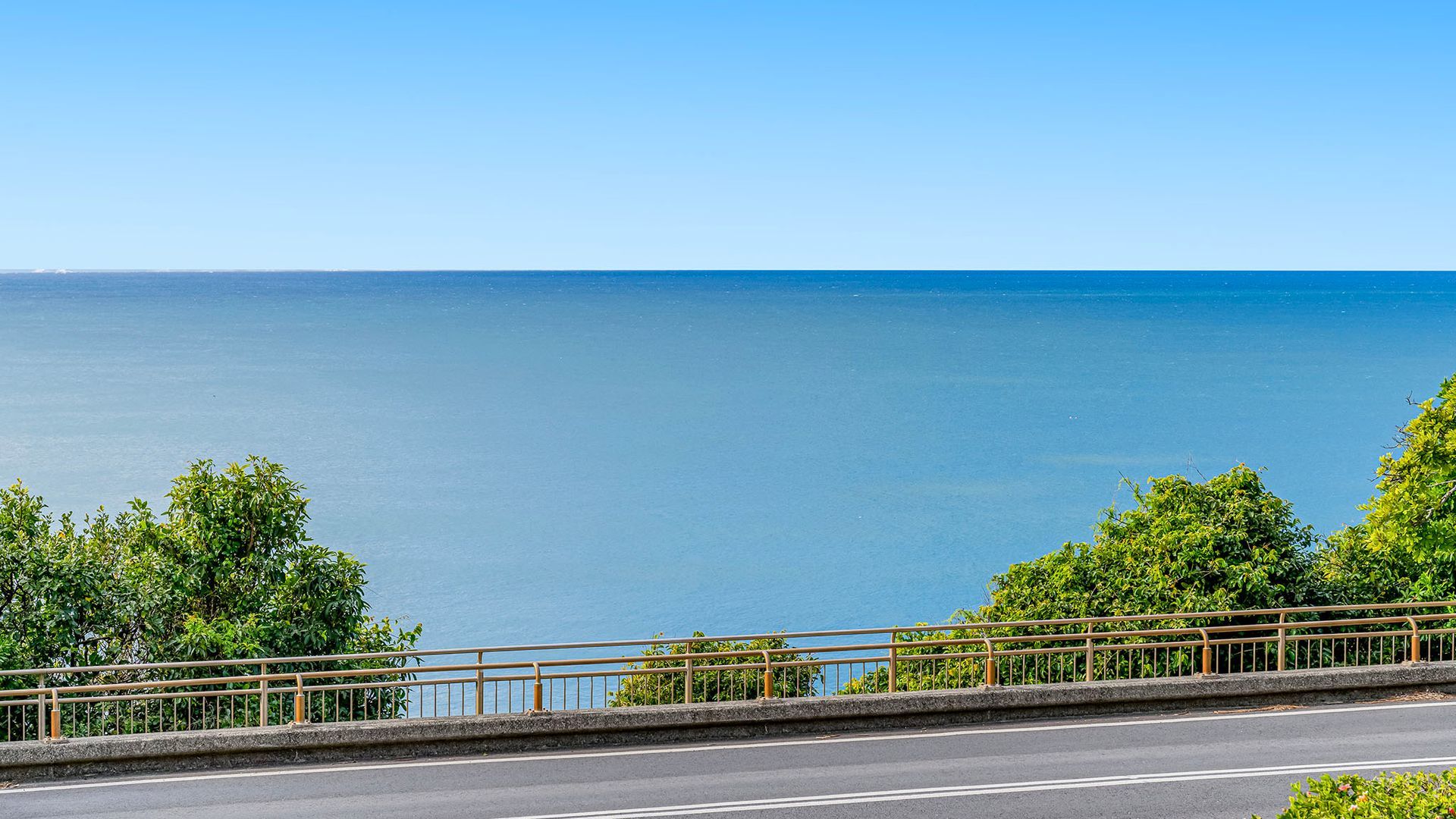 A coastal road with a metal guardrail, trees, and a clear view of the sea.