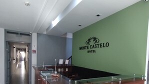 A hotel reception area with a wooden reception desk, a green wall with the hotel name, and a corridor leading to other rooms. - HOTEL MONTE CASTELO LTDA. (Rio de Janeiro)