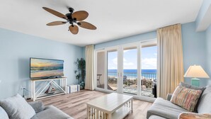 Uma sala de estar ampla com uma grande janela que oferece vista para a praia, um ventilador de teto e uma televisão sobre um suporte.