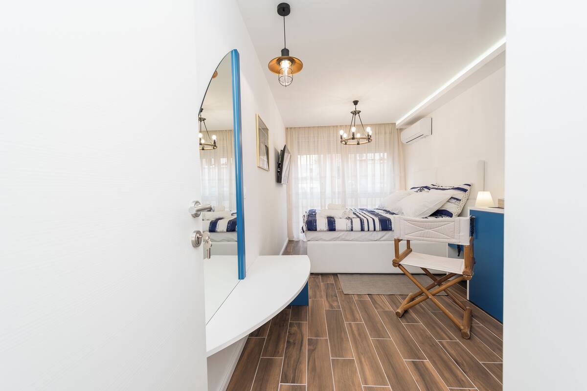 A modern hotel room with a white vanity, a large mirror, a wooden floor, a bed with a striped blanket, and a blue accent wall.