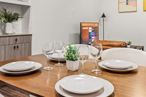 A dining table set with white plates and wine glasses, a small potted plant, and a television displaying a streaming service interface. - Classy Modern Studio with Pool plus Golf Sim (Austin)