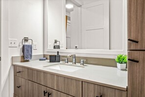 A modern bathroom with a white sink, two faucets, a mirror, and wooden cabinets. - Classy Modern Studio with Pool plus Golf Sim (Austin)