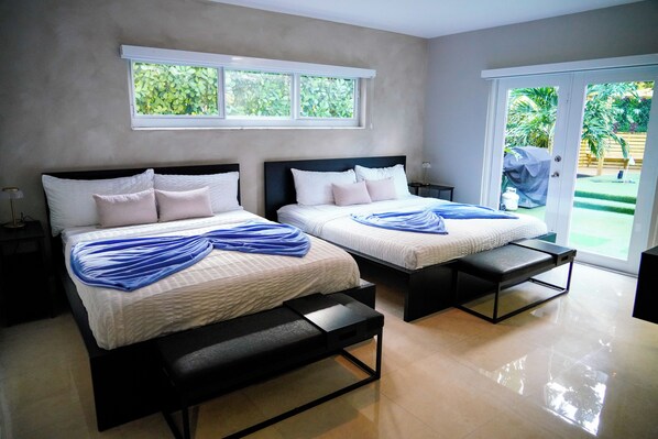 A bedroom with two beds, a bench, a nightstand, and a window with a view of greenery.