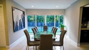 Un coin repas avec une table en verre et quatre chaises, une grande fenêtre donnant sur un paysage verdoyant et un tableau encadré au mur.