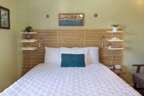 A neatly made bed with a white quilt, a teal pillow, and wooden slats on the headboard. There are shelves with plants and books, and a painting above the headboard.