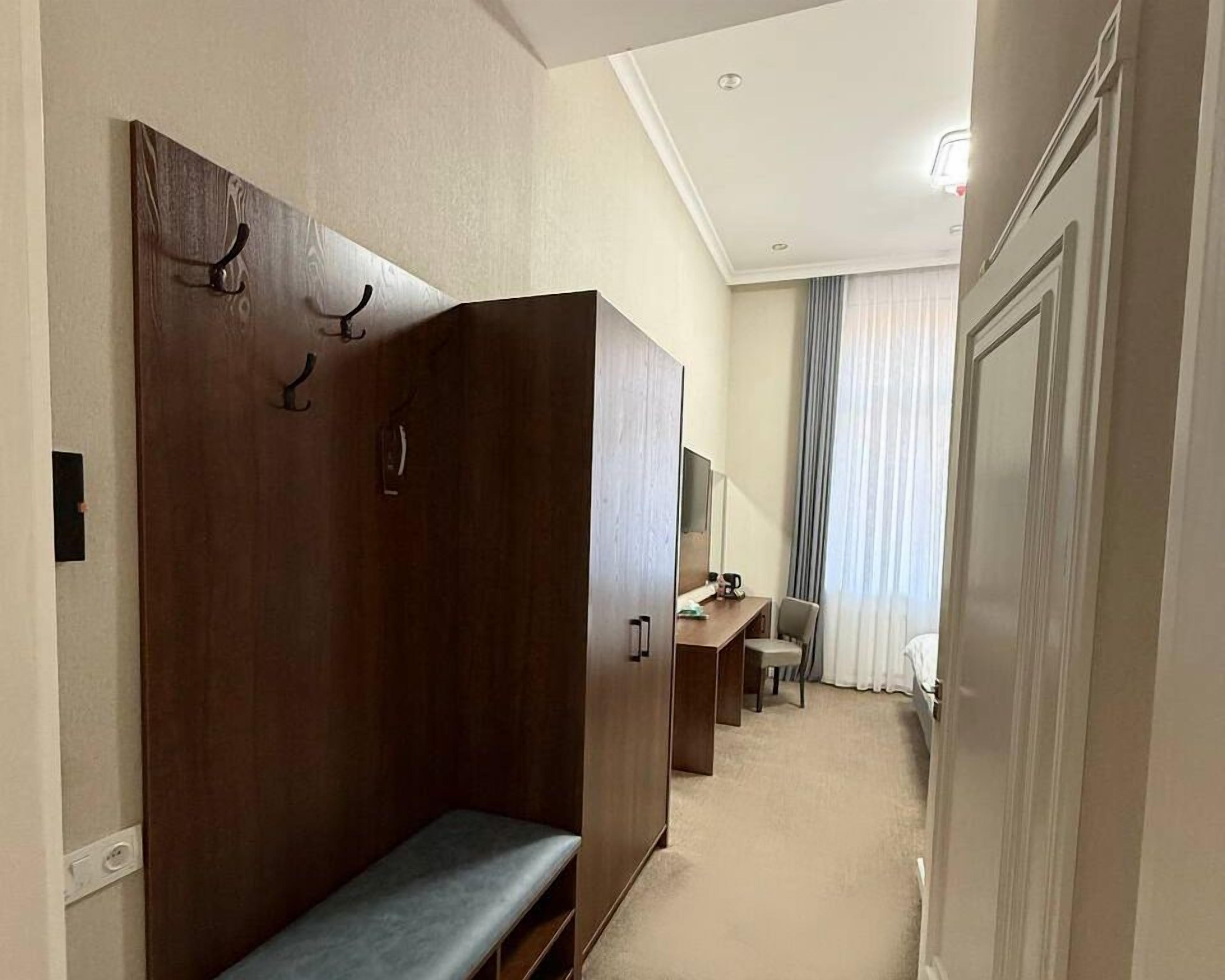 A hotel room with a wooden wardrobe, a bench, a desk with a chair, and a large window with curtains.