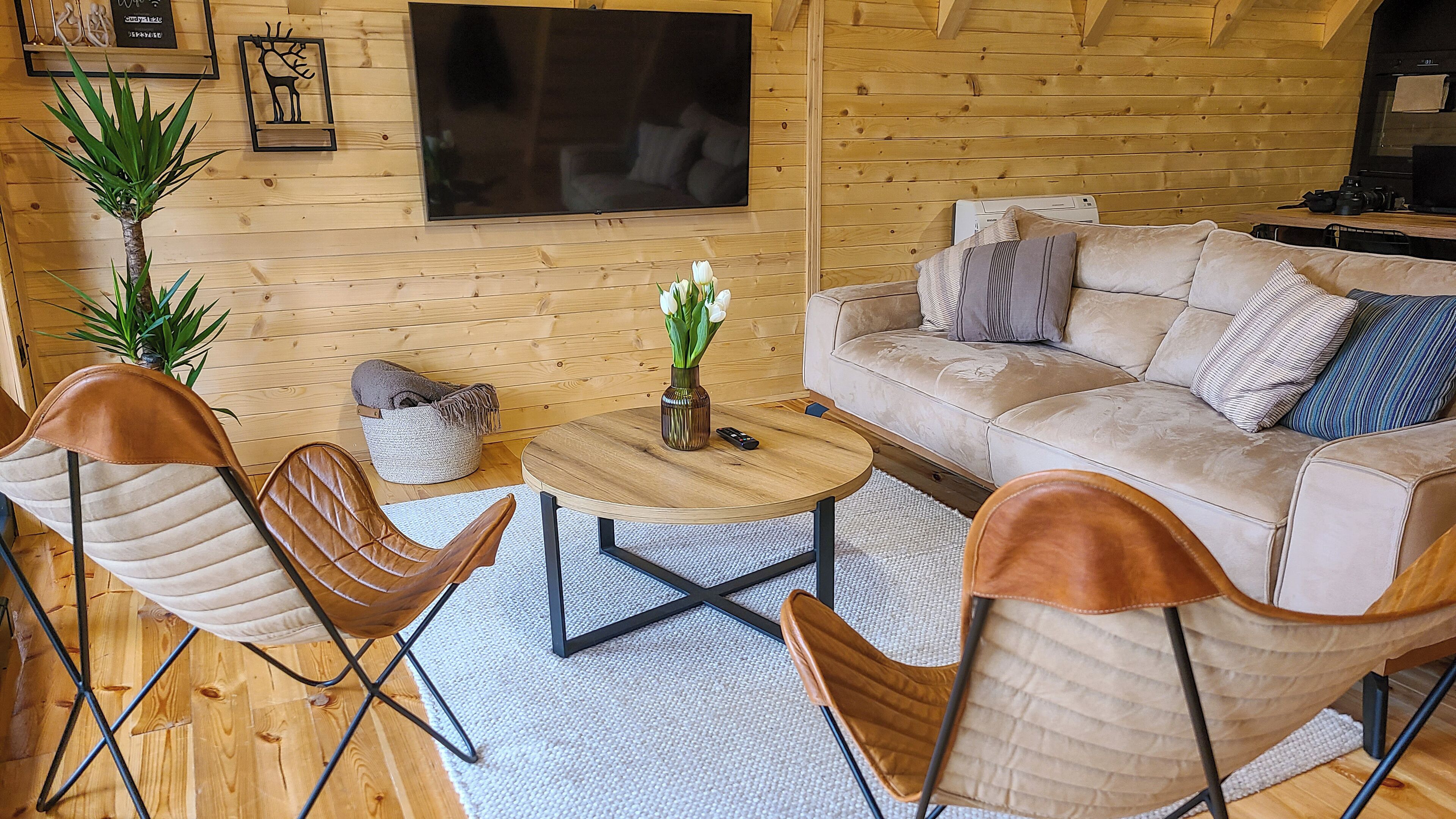 Een gezellige woonkamer met een houten inrichting, een beige bank, twee leren fauteuils, een salontafel en een flatscreen televisie aan de muur.