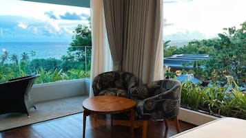 Une chambre d’hôtel avec un grand lit, une petite table ronde, deux fauteuils à motifs et une vue sur l’océan et la verdure.