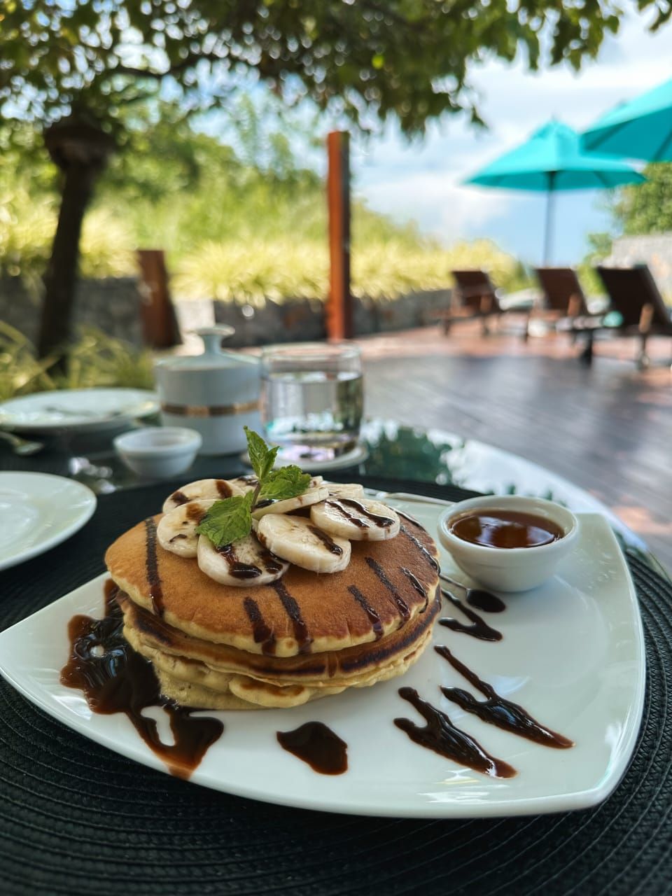 A breakfast setting with pancakes topped with banana slices and a mint leaf, served on a white plate with chocolate syrup.