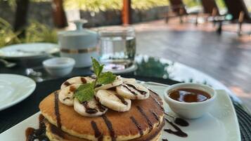 A breakfast setting with pancakes topped with banana slices and a mint leaf, served on a white plate with chocolate syrup.