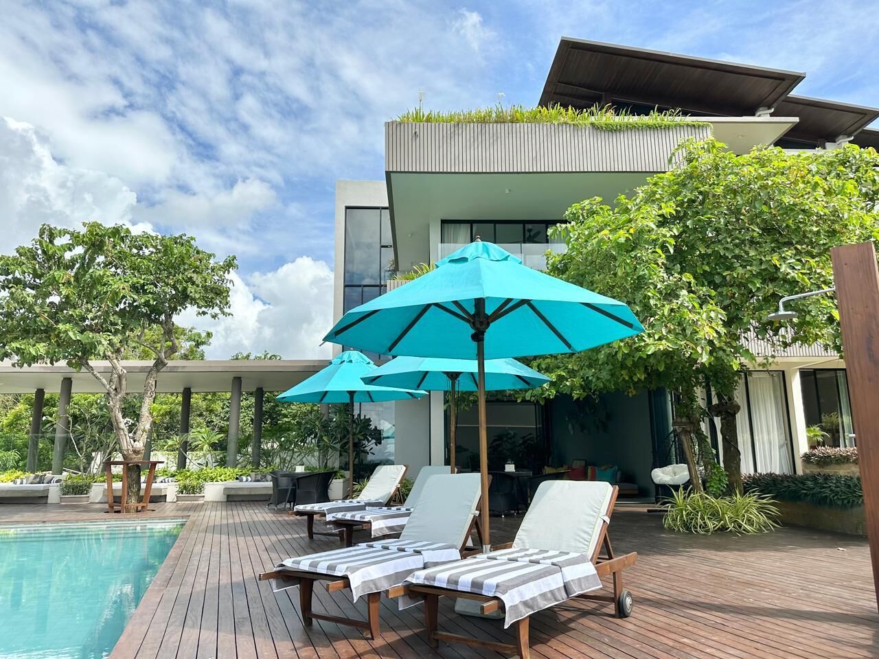 Un espace piscine extérieur moderne, doté de chaises longues et de parasols, entouré de verdure et bordé d’un bâtiment contemporain.