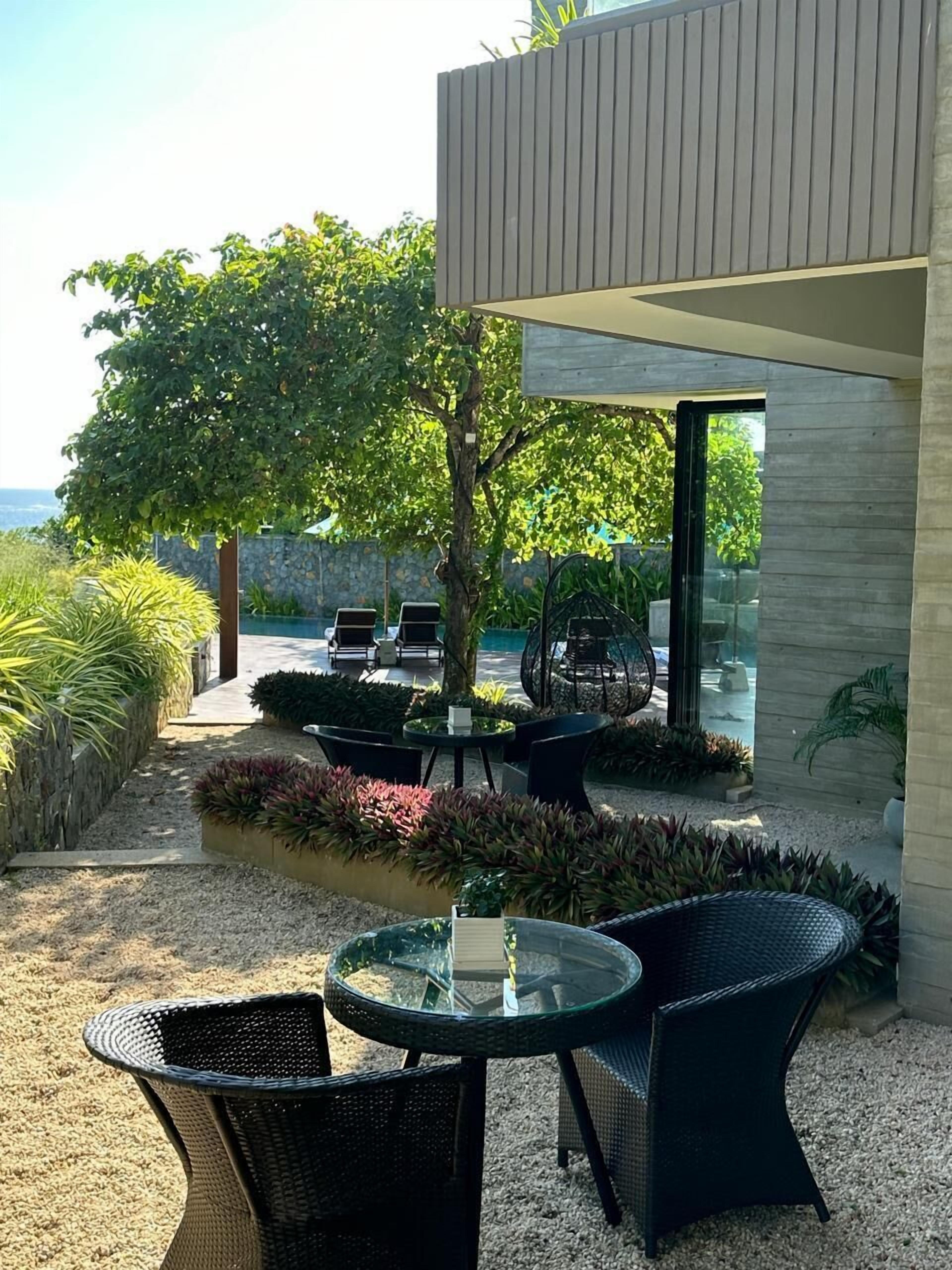 A modern outdoor patio with a glass table and chairs, surrounded by lush greenery and a view of the sea.