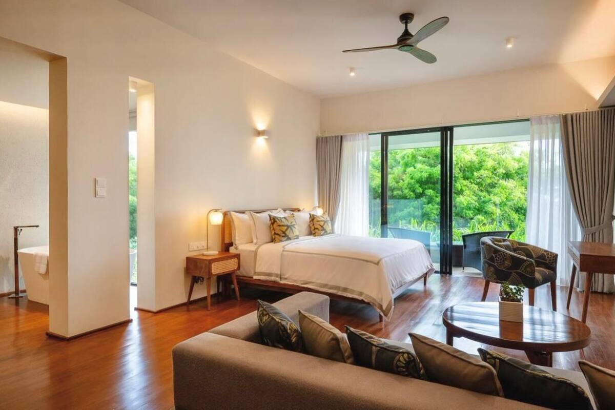 A spacious bedroom with a large bed, a sitting area with a sofa and a table, and a balcony with a view of greenery.