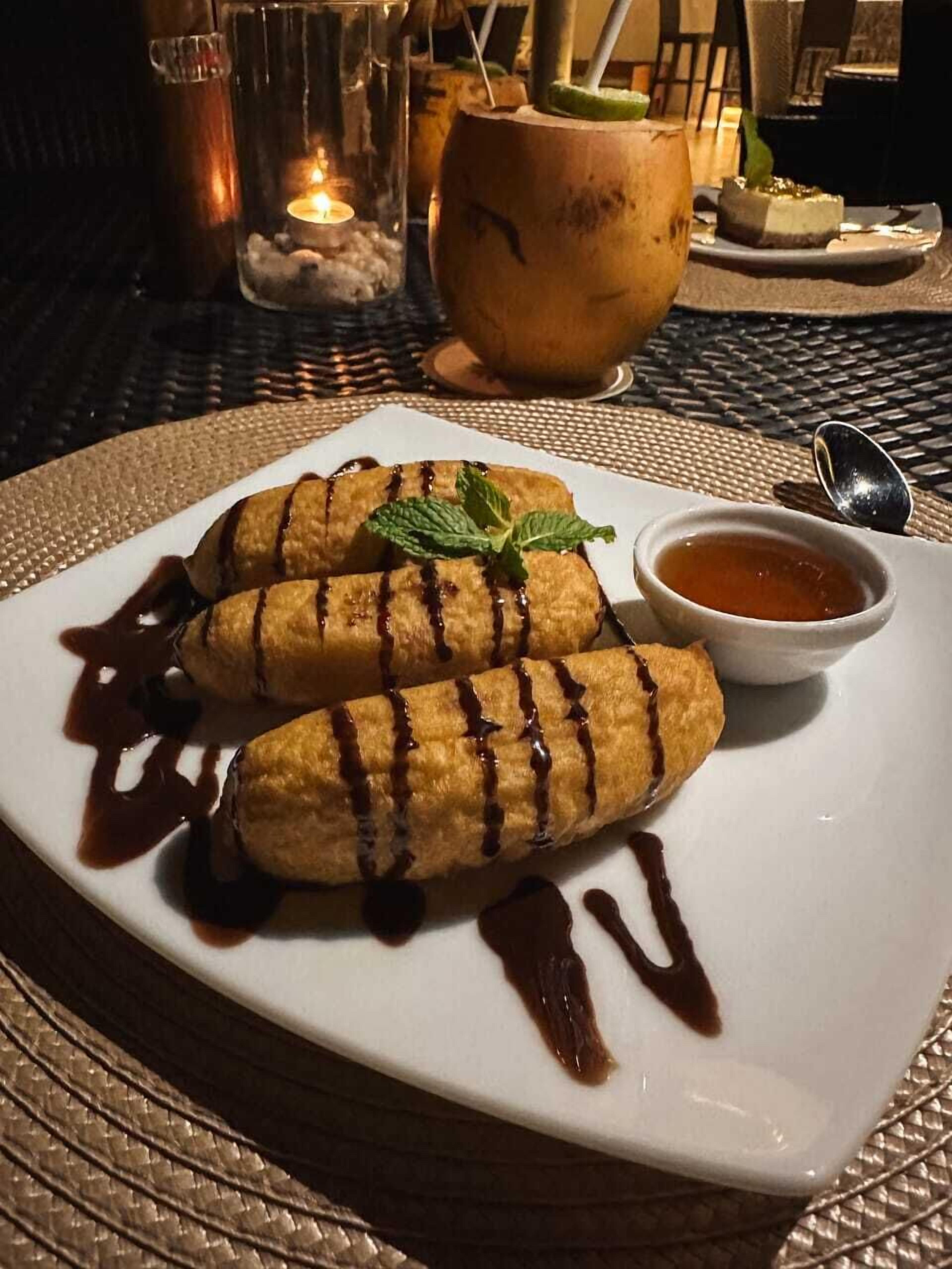 A plate of banana fritters with chocolate sauce, a small bowl of sauce, and a drink in the background.