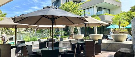 An outdoor patio with tables and chairs, umbrellas, and a modern building in the background.