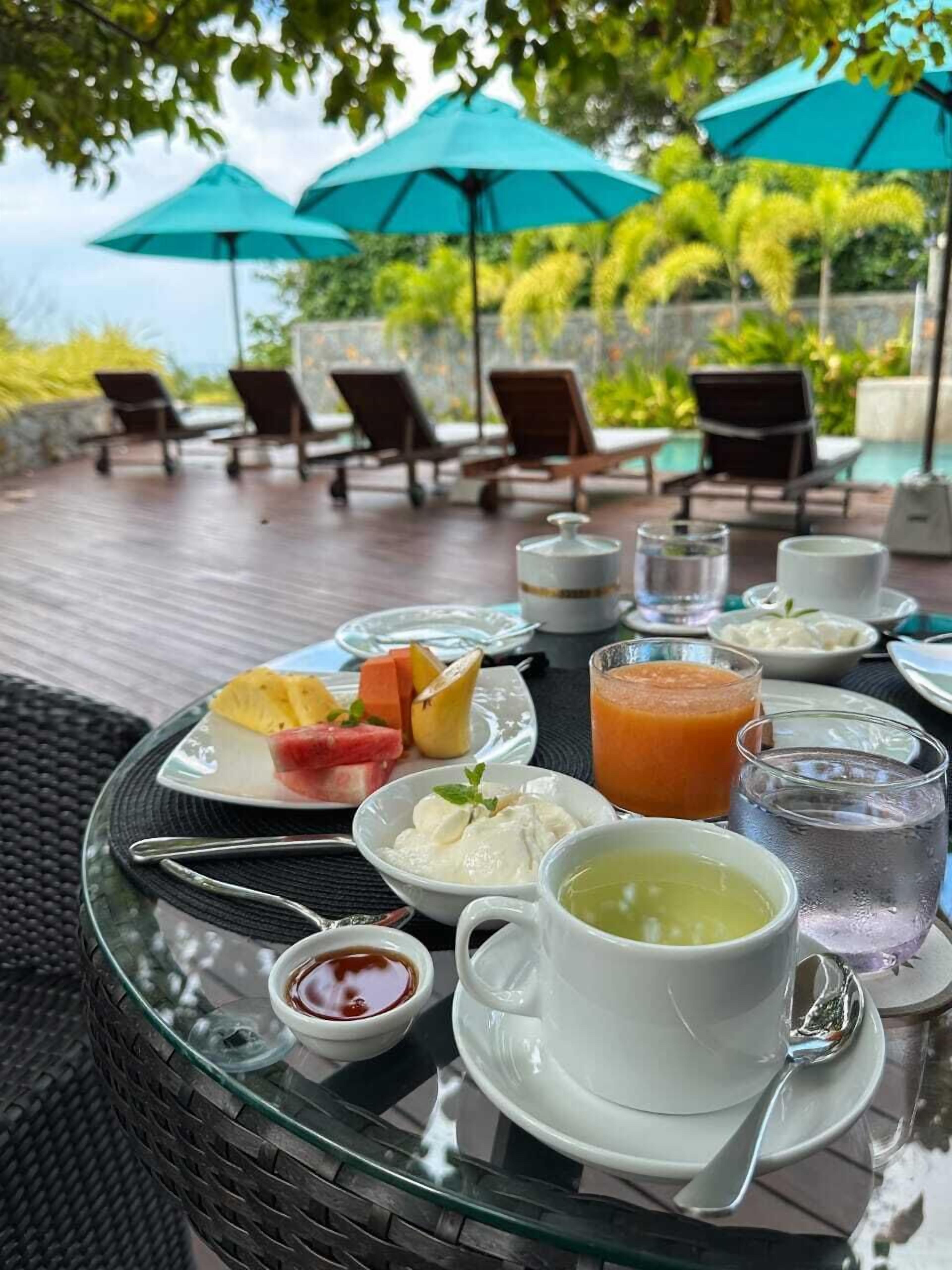 Una mesa servida con diversos alimentos y bebidas, entre ellos frutas, un batido y una taza de té, bajo un paraguas de vidrio en un patio al aire libre.
