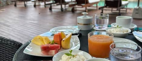 Una mesa servida con diversos alimentos y bebidas, entre ellos frutas, un batido y una taza de té, bajo un paraguas de vidrio en un patio al aire libre.