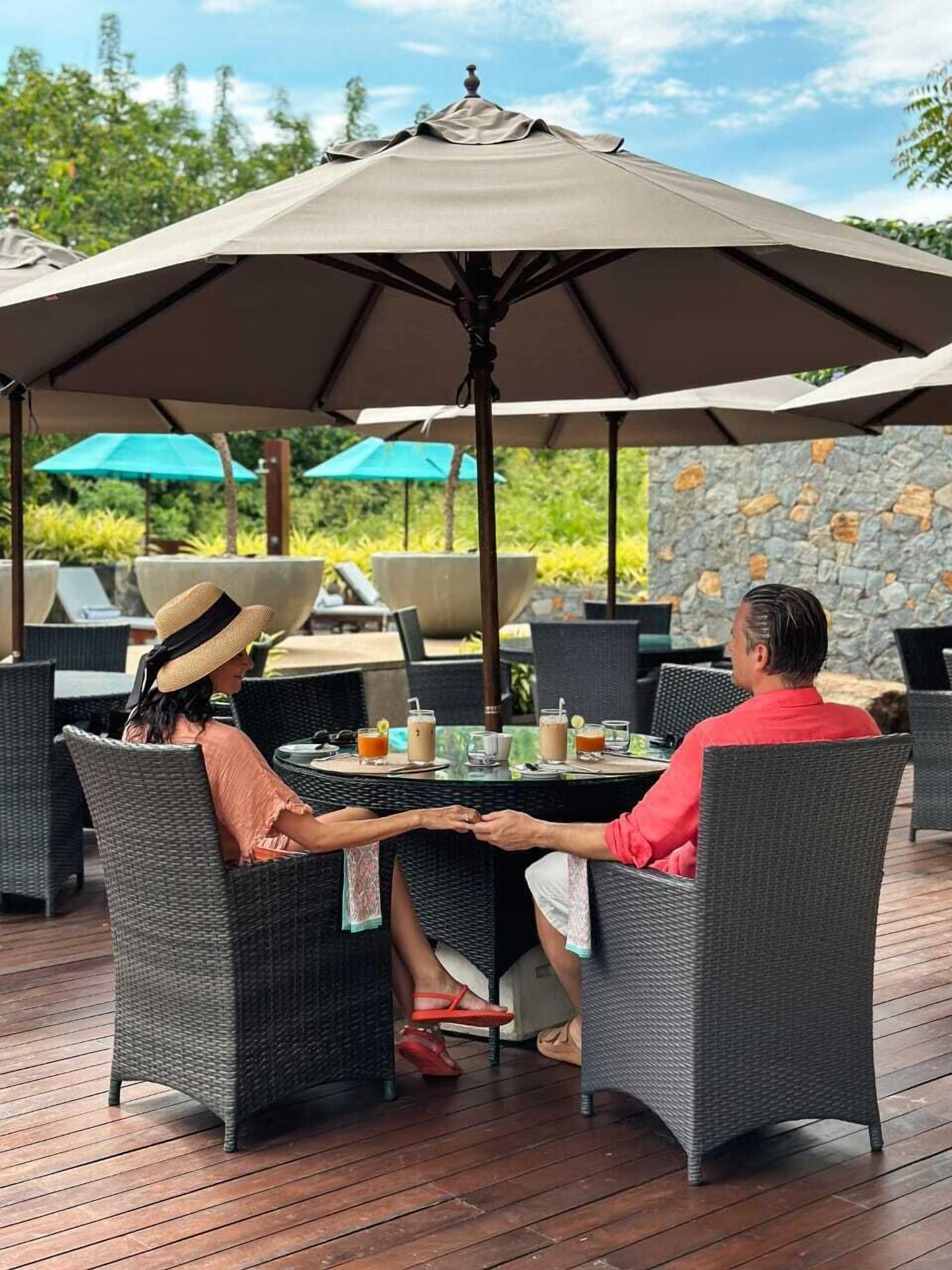 A couple seated at an outdoor table with a large umbrella, surrounded by wicker chairs and a stone wall.