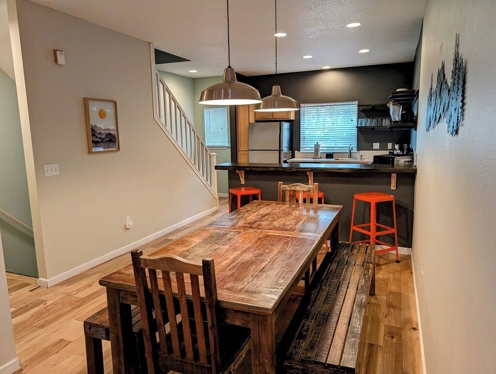 A dining area with a wooden table and benches, a kitchen with a black countertop and red stools, and a staircase leading upstairs.