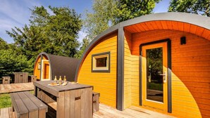 Een klein, geel, halfrond houten gebouwtje met een deur en raam, omgeven door een houten terras met picknicktafels.