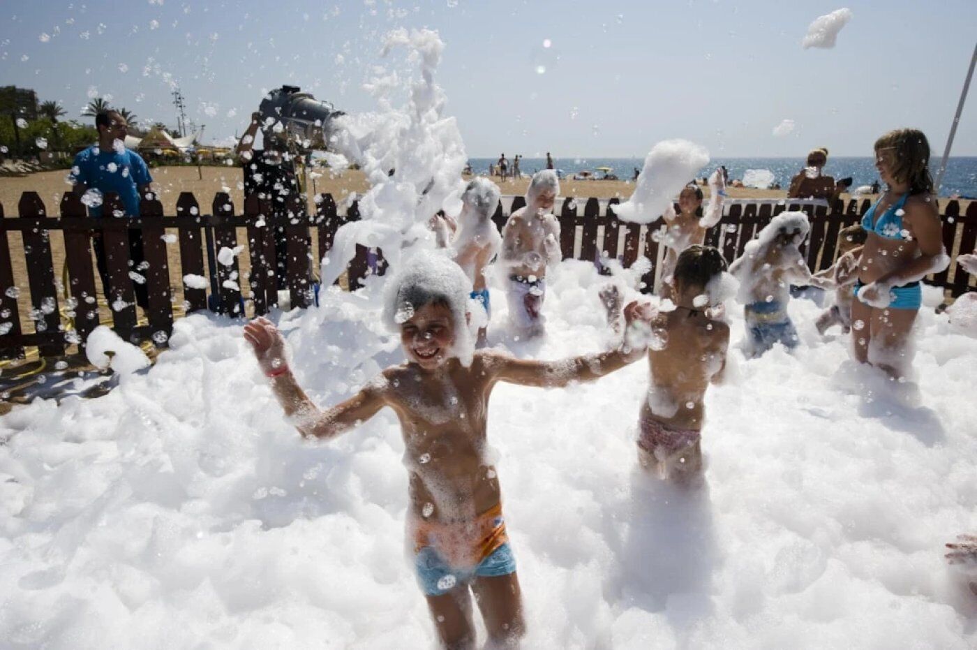 Niños jugando en la espuma de la playa, con una cerca de madera y gente al fondo.