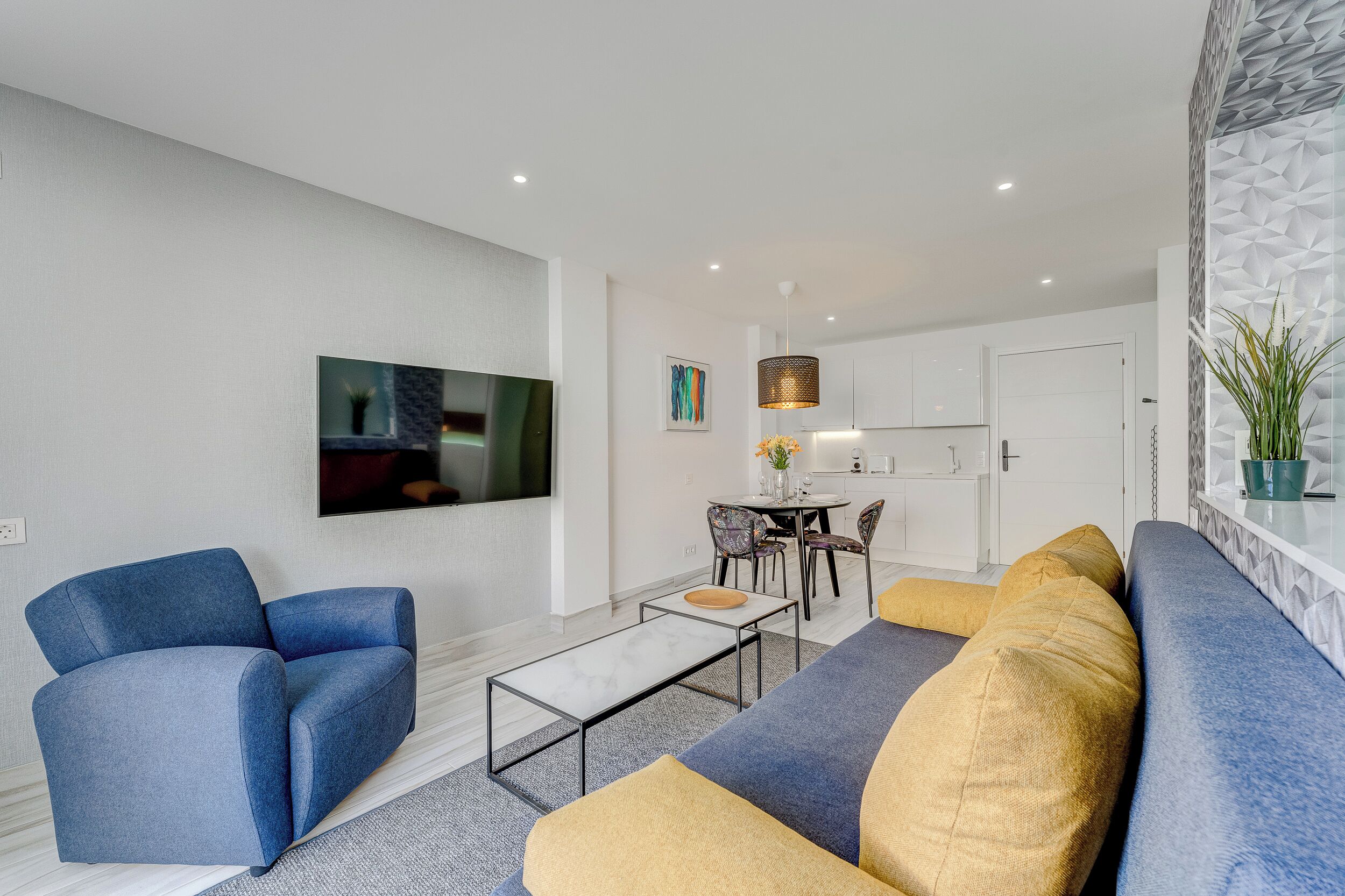Un salon moderne avec un canapé, un fauteuil et une table basse. On y trouve également un coin repas avec des chaises et une table, ainsi qu’une cuisine à l’arrière-plan.