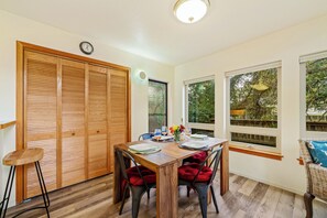 Un comedor con una mesa de madera dispuesta para cuatro personas, sillas con cojines rojos, un reloj en la pared y ventanas amplias con vistas a árboles.