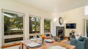Un comedor con una mesa de madera dispuesta para cuatro personas, ventanas amplias, una chimenea y un televisor de pantalla plana montado en la pared.