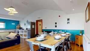 Un comedor con una mesa de madera dispuesta para seis personas, rodeada de sillas de metal. Se ve, al fondo, una zona de cocina con un refrigerador y un lavamanos.