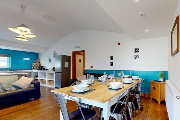 A dining area with a wooden table set for six, surrounded by metal chairs. A kitchen area with a refrigerator and a sink is visible in the background.