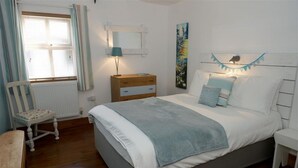 Un dormitorio con cama, mesita de noche, cómoda, silla, ventana con cortinas y lámpara de pared.