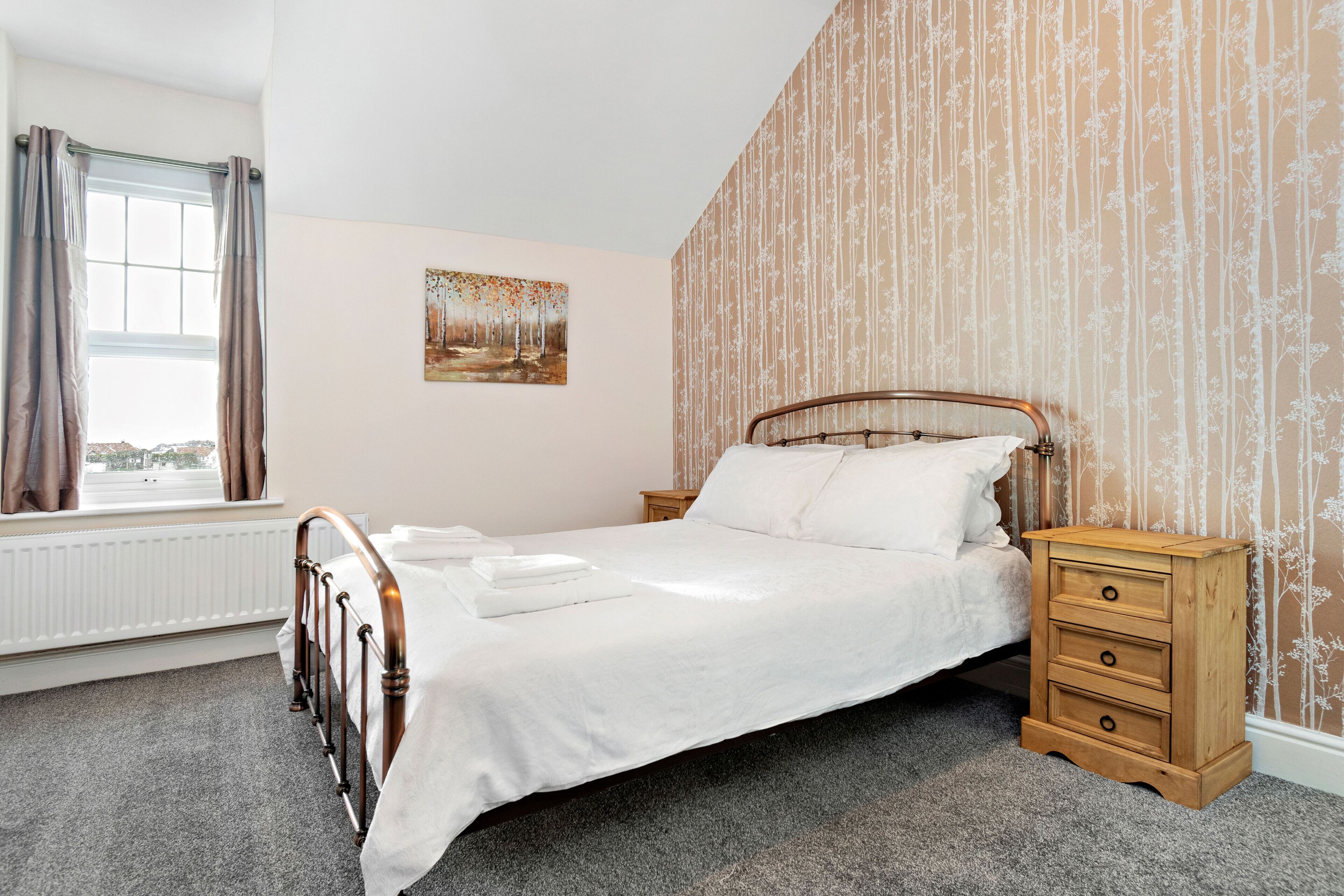 Un lit bien fait, avec un cadre en bois et du linge de lit blanc, une table de chevet en bois avec des tiroirs, un radiateur, une fenêtre avec des rideaux et un mur recouvert d’un papier peint à motifs floraux verticaux.