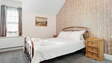 Un lit bien fait, avec un cadre en bois et du linge de lit blanc, une table de chevet en bois avec des tiroirs, un radiateur, une fenêtre avec des rideaux et un mur recouvert d’un papier peint à motifs floraux verticaux.
