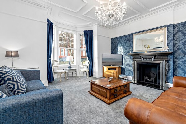 Ein Wohnzimmer mit blauem, gemustertem Wandpapier, einem Kamin, einem Fernseher und einer Chandelier.