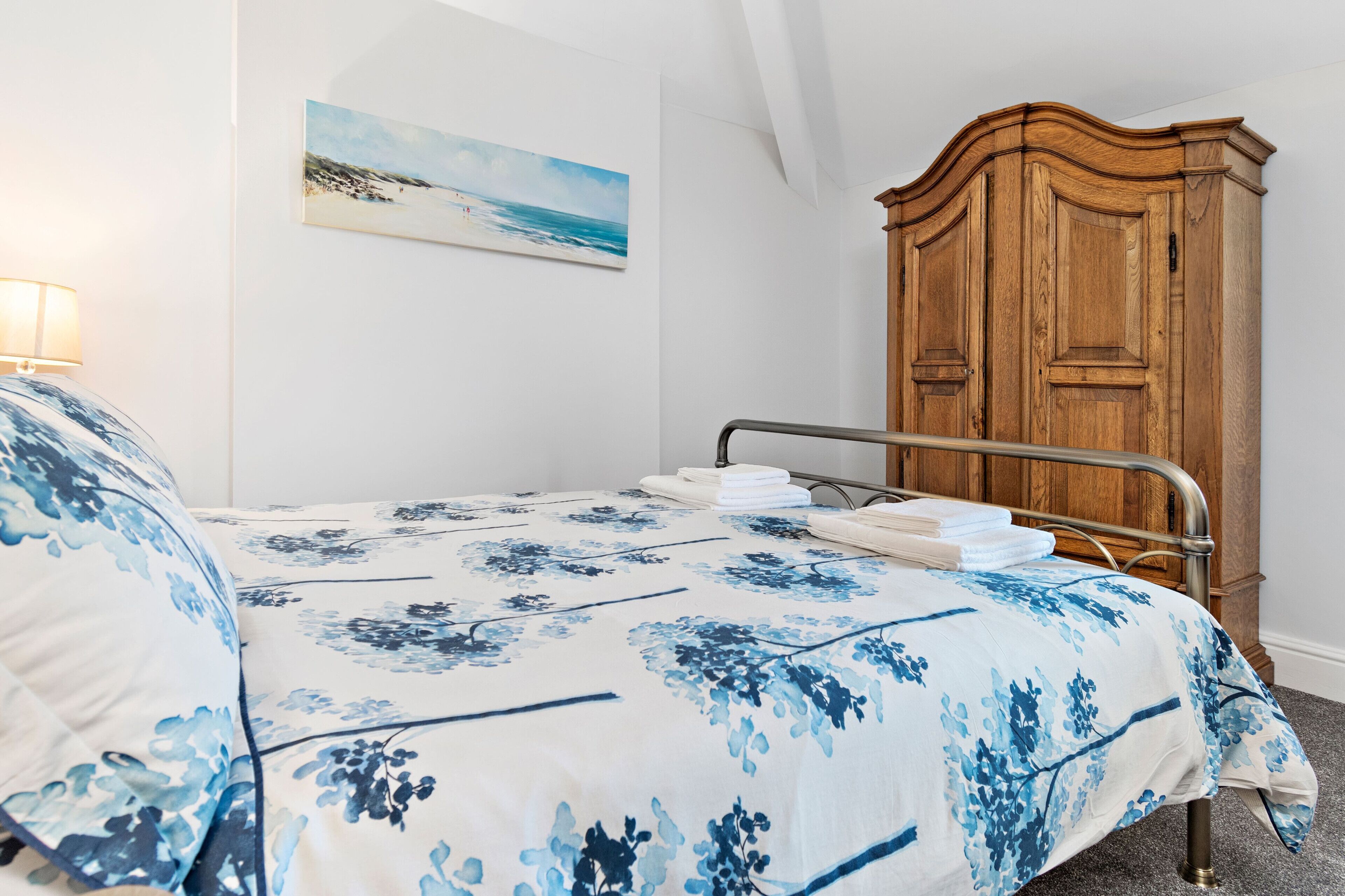 A bedroom with a bed, a wooden wardrobe, a painting of a beach, and a lamp.