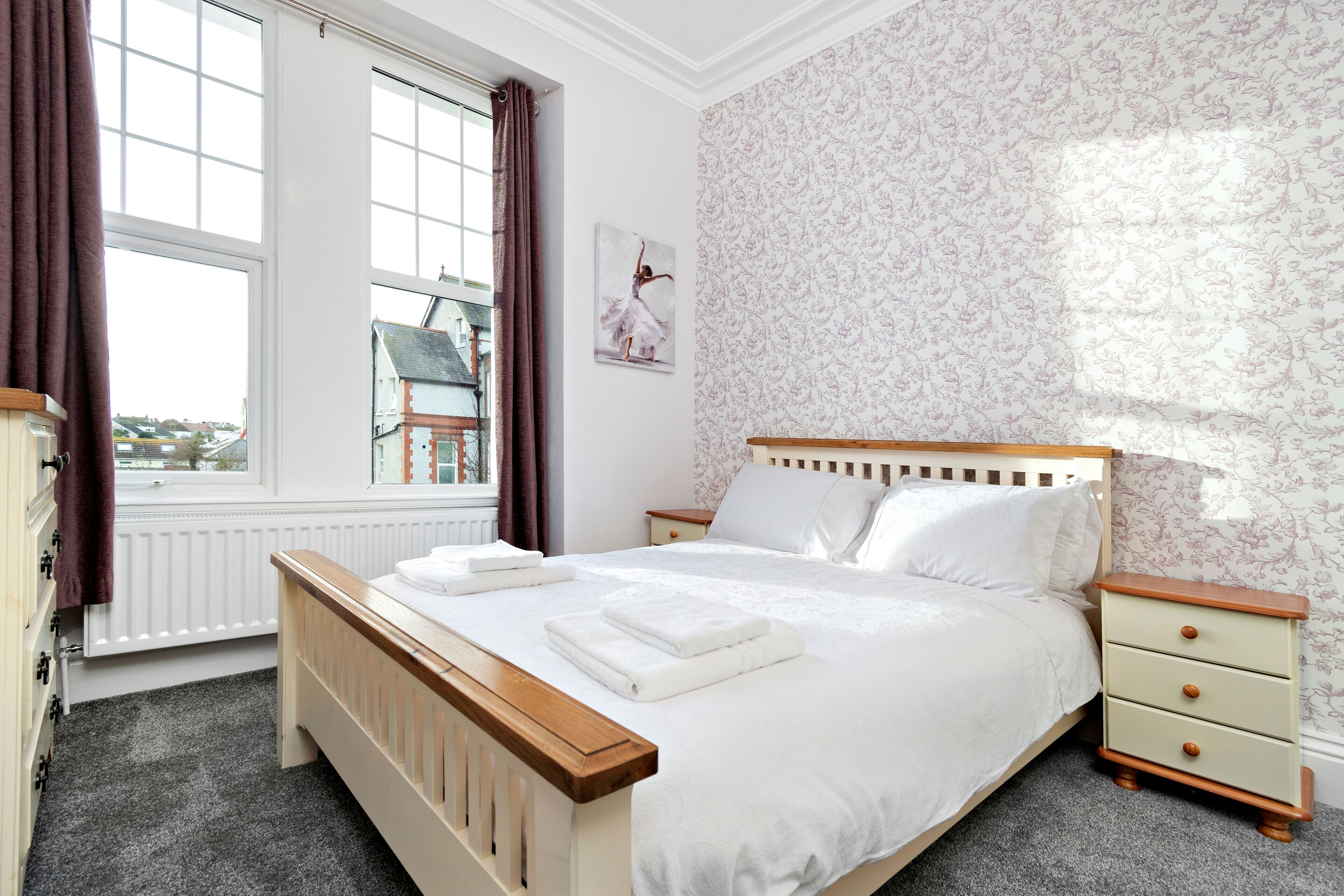 Un dormitorio con un ventanal grande, una cama con ropa blanca, un cabecero de madera, una mesita de noche y un papel tapiz floral en la pared.